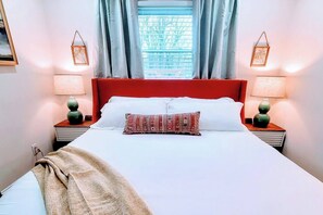Une chambre d’hôtel avec un grand lit, deux tables de chevet avec des lampes, une fenêtre avec des rideaux et une vue sur les arbres à l’extérieur.