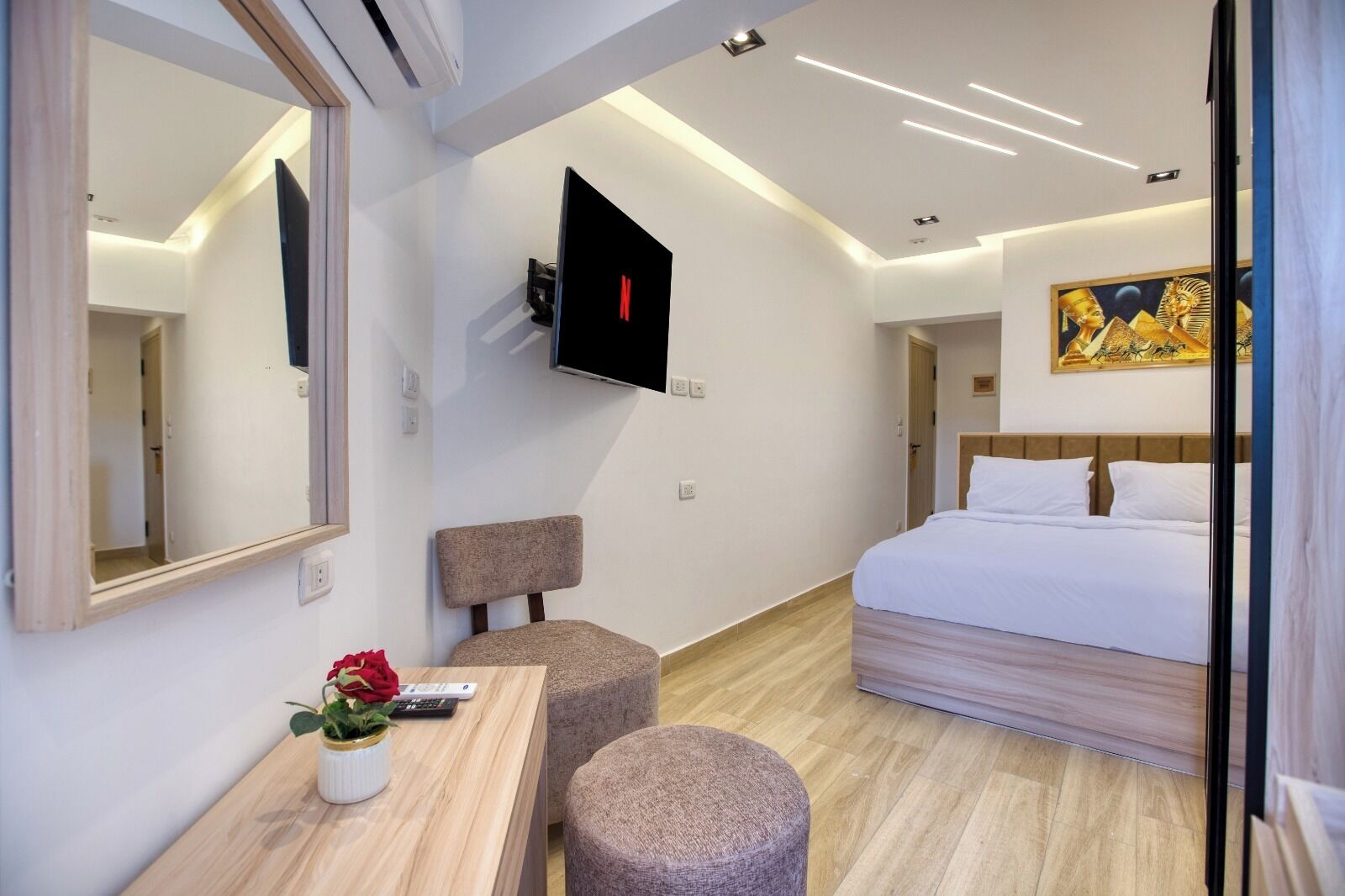 Ein modernes Hotelzimmer mit einem Bett, einem an der Wand montierten Fernseher, einem kleinen Tisch mit Blumenvase und einer Holzbank.