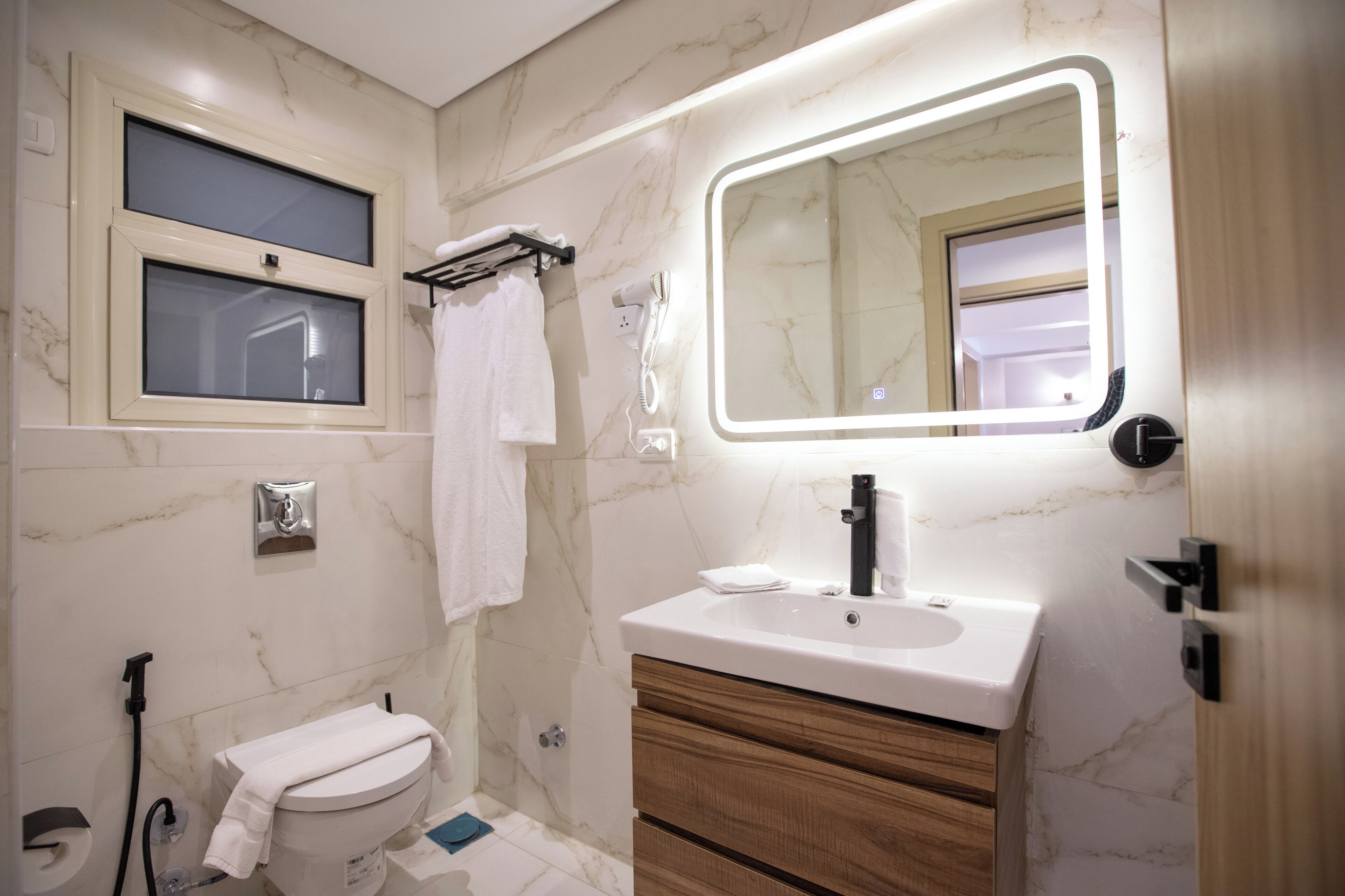 A bathroom with a toilet, sink, vanity, mirror, towel rack, and a window.