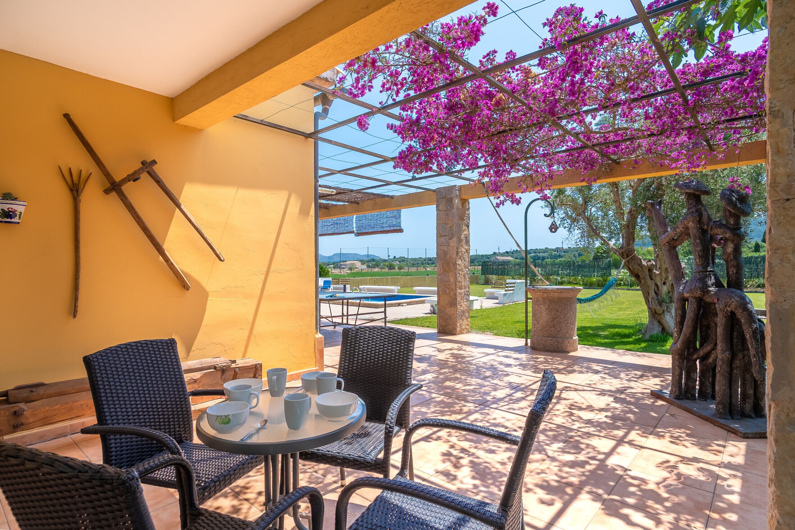 An outdoor patio with wicker furniture, a table with cups and saucers, and a view of a pool and greenery.
