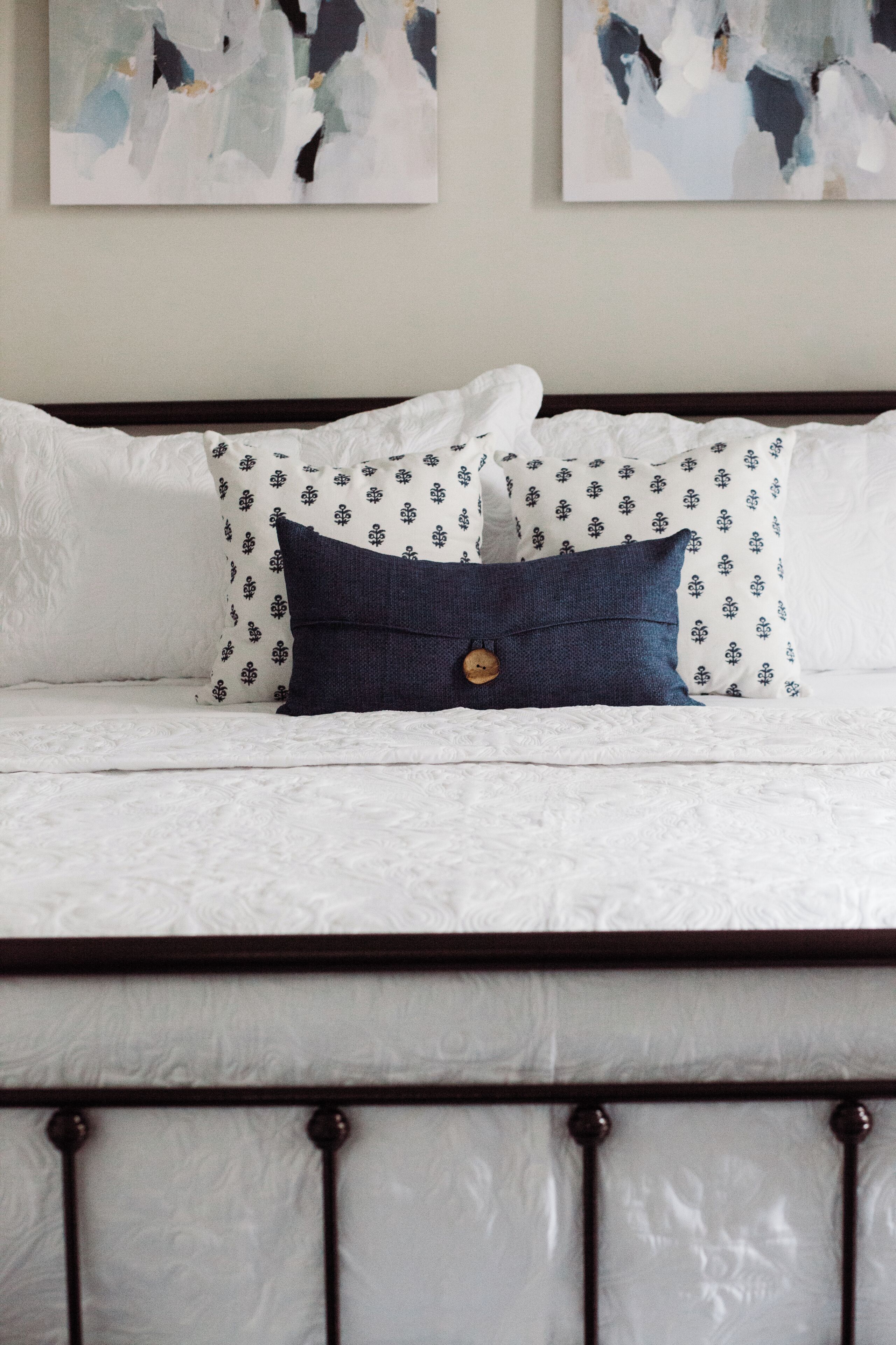 Un lit avec du linge de lit blanc et des coussins à motifs, une tête de lit en bois et une œuvre d’art abstraite accrochée au mur.
