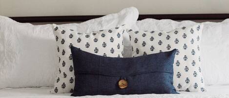 Un lit avec du linge de lit blanc et des coussins à motifs, une tête de lit en bois et une œuvre d’art abstraite accrochée au mur.