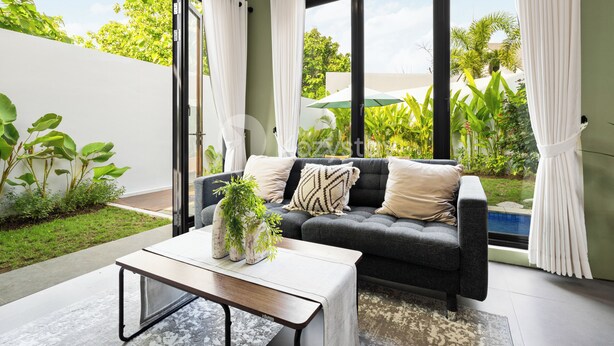 A modern living room with a grey sofa, a coffee table, and a view of a swimming pool and greenery outside. - Marea Villa by Kozystay - Jimbaran (Jimbaran)
