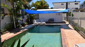 A rectangular swimming pool with a brick border, surrounded by a patio area with white lounge chairs and a blue shade sail.
