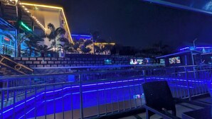 Vista noturna de um bar no terraço iluminado com luzes de neon azuis, palmeiras e uma escadaria que leva a um nível superior.