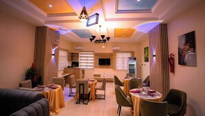 Uma sala de jantar de hotel com mesas, cadeiras, um lustre e uma televisão.