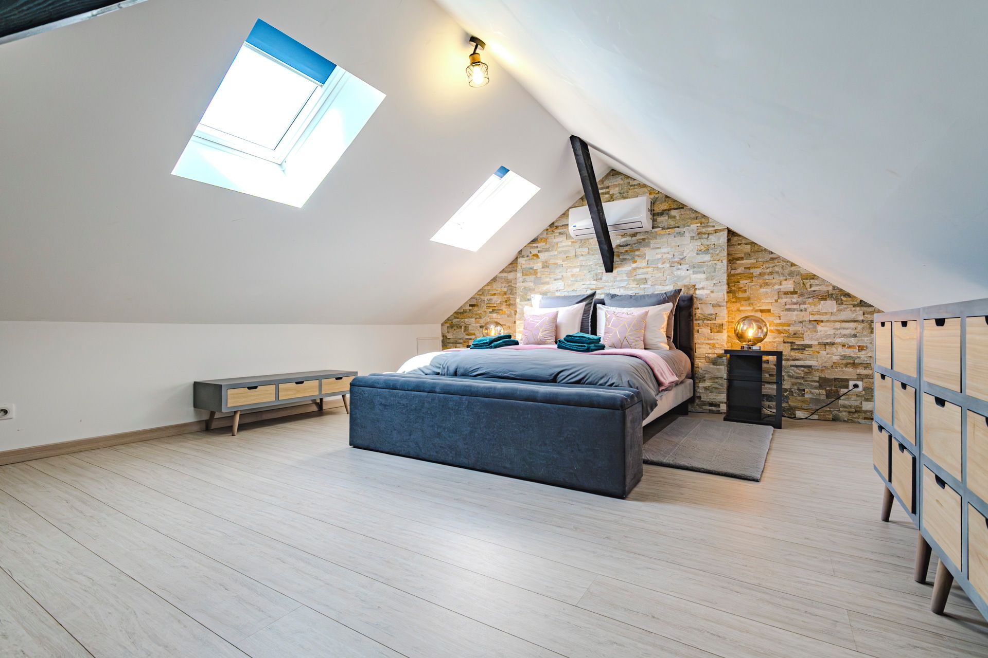 Un dormitorio con techo inclinado, una cama con almohadas y una colcha, una mesita de noche con lámpara, y una ventana.