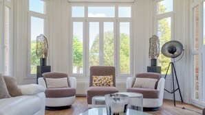 A modern living room with two white sofas, a brown armchair, and a glass coffee table. There are large windows with a view of trees and a brick wall.