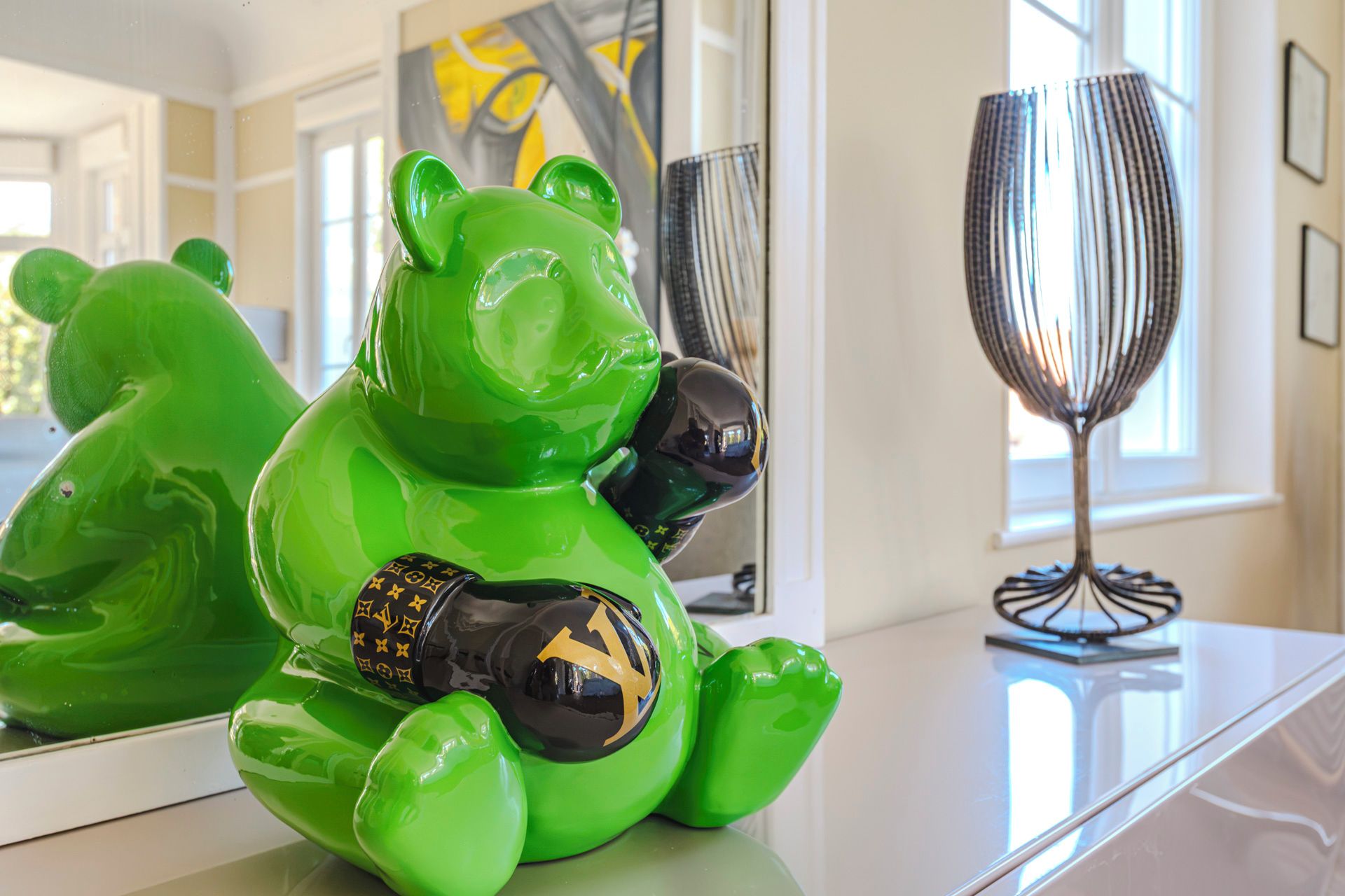 A green bear figurine with a Louis Vuitton bag on a white surface, a metallic vase, and a window with a view.