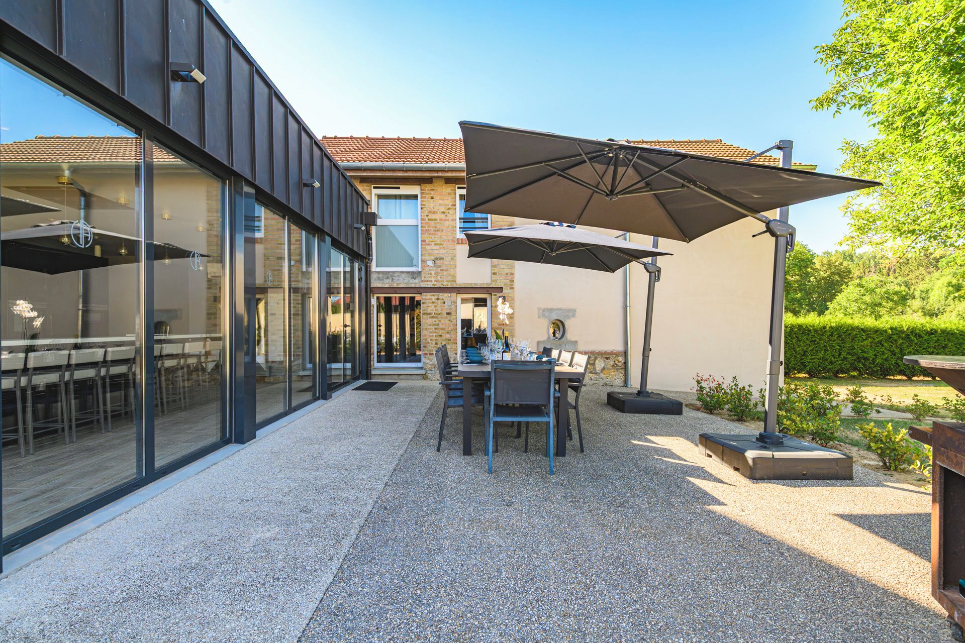 Et moderne udendørs spiseområde med et bord, stole og en stor parasol, beliggende på en grusbelagt terrasse.