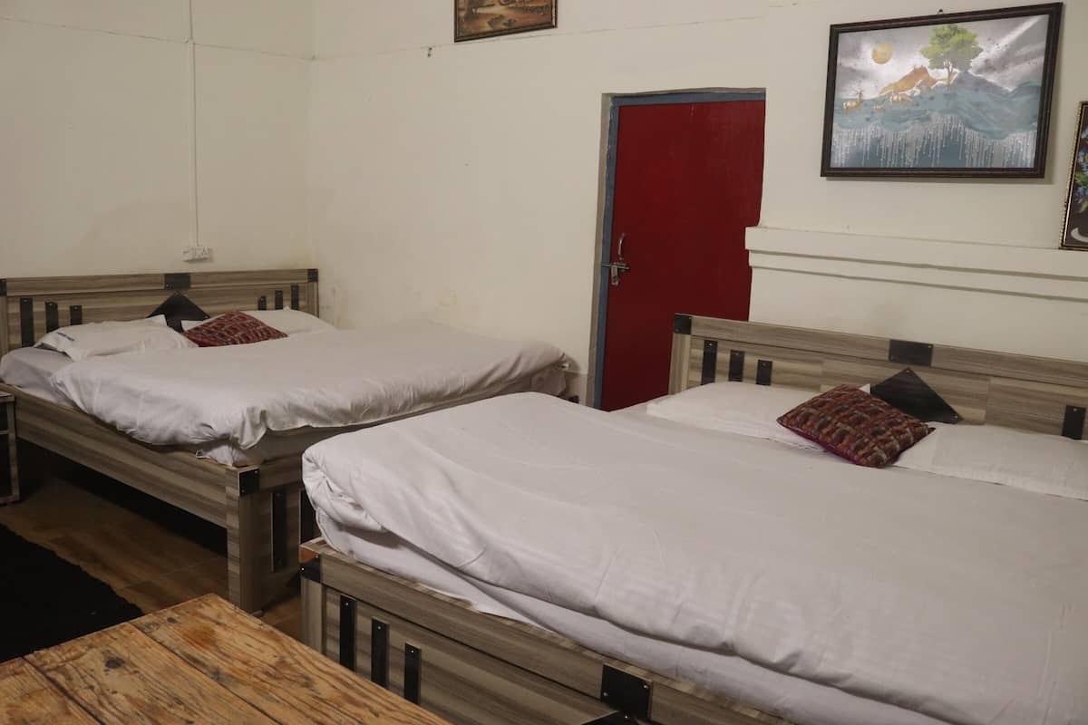 Una habitación con dos camas de madera, cada una con ropa de cama blanca y una almohada estampada. Se ve una puerta roja al fondo, y un cuadro enmarcado cuelga en la pared.