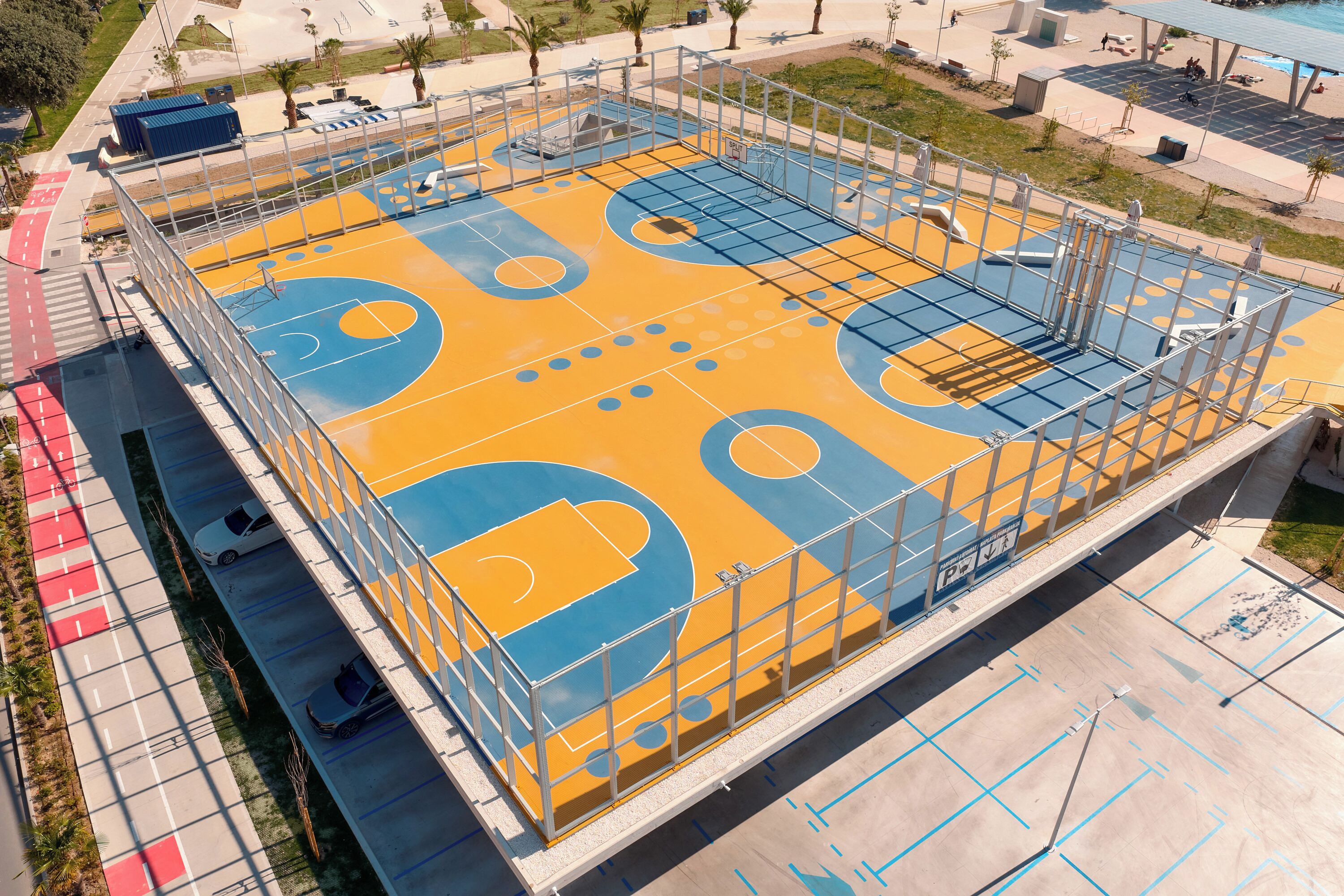 A basketball court with a metal fence, a parking area, and a pedestrian walkway.