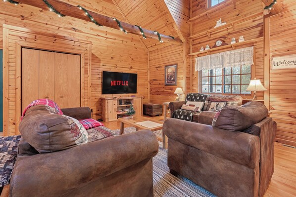 A cozy log cabin interior with a wooden TV stand, two brown sofas, a coffee table, and a window with a view.
