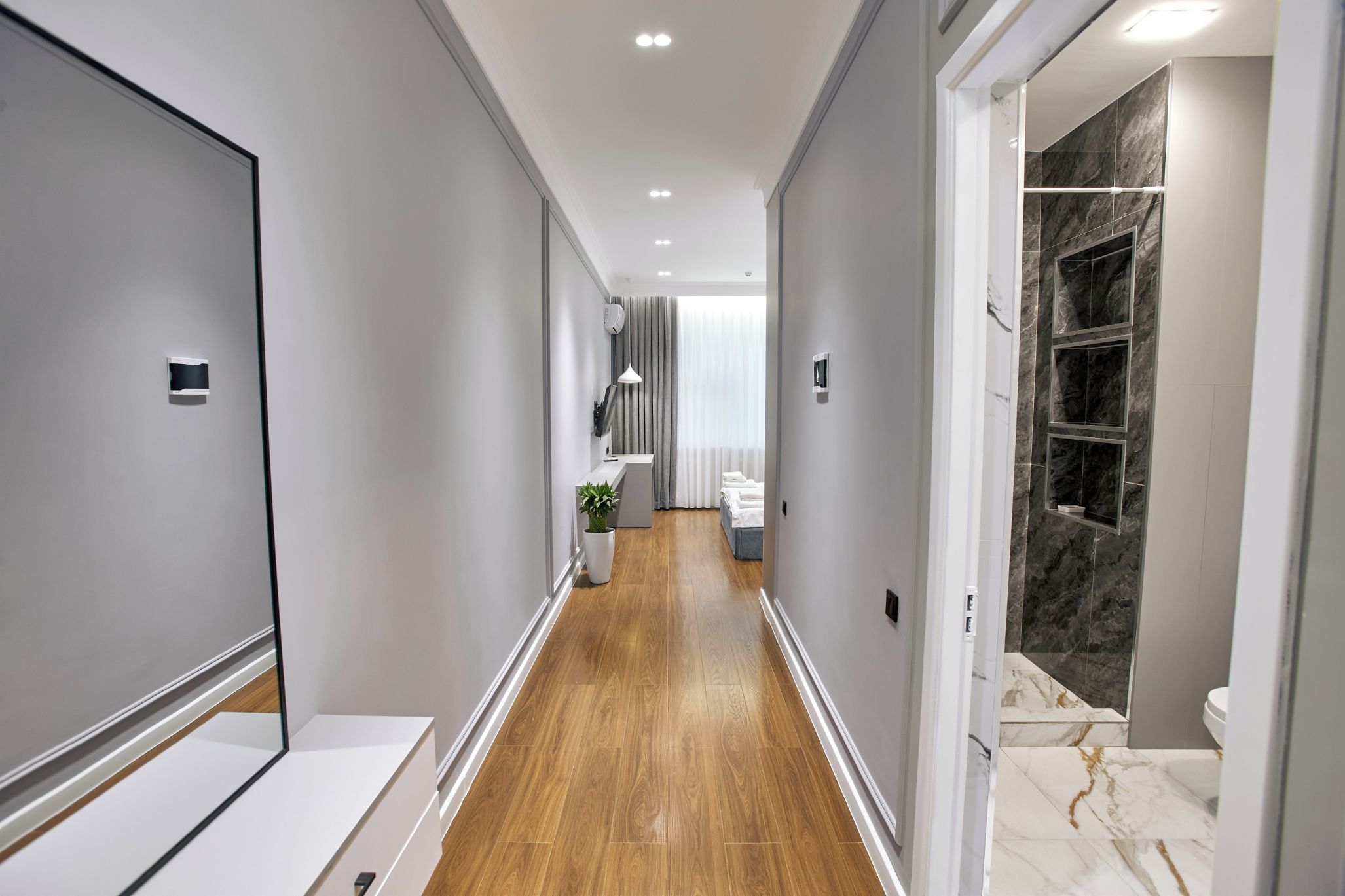 Un couloir d’hôtel moderne avec un plancher en bois, un lit recouvert de draps blancs, un bureau avec une lampe et une salle de bain dotée d’une douche en marbre.