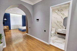 A modern hotel room with a wooden floor, a bed with blue bedding, a small desk with a lamp, and a bathroom with marble tiles.