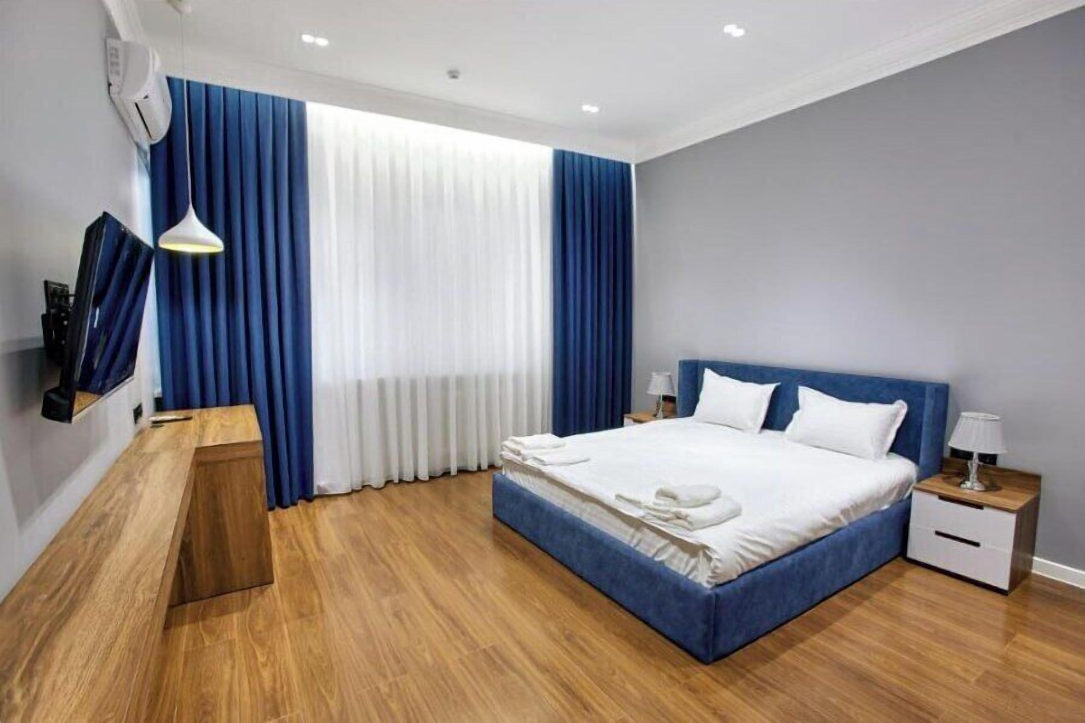 A modern hotel room with a large bed, a wooden desk, a nightstand, and a TV mounted on the wall.