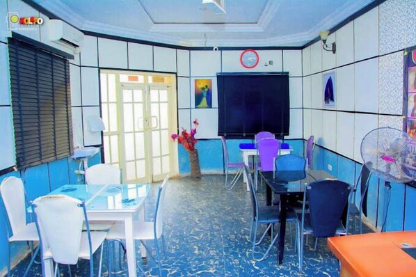 Une salle de conférence équipée d’un grand écran, de chaises blanches et bleues, d’une table et d’une horloge murale.