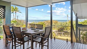 A covered outdoor dining area with a view of the ocean, wooden furniture, and a clear sky. - Caloundra Retro Beach House in Front of Ocean (Caloundra Region)