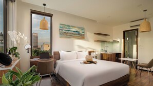 A modern hotel room with a large bed, a desk with a laptop, a small table, and a chair. There is a window with a view of buildings and a painting on the wall. - Dragon View Riverfront Hotel  (Da Nang)