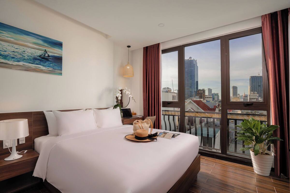 Habitació d'hotel amb un llit gran, vista a la ciutat, un quadre a la paret i una planta en test.