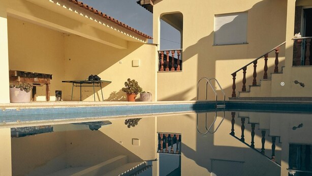 Une piscine dont l’eau est si claire qu’elle reflète parfaitement l’environnement, un patio couvert et un escalier menant à un balcon.
