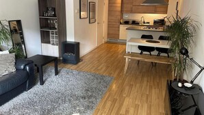 Un salón moderno con alfombra gris, sofá de cuero negro y mesa de comedor de madera con sillas negras. Se ve una cocina con armarios de madera y estufa al fondo.