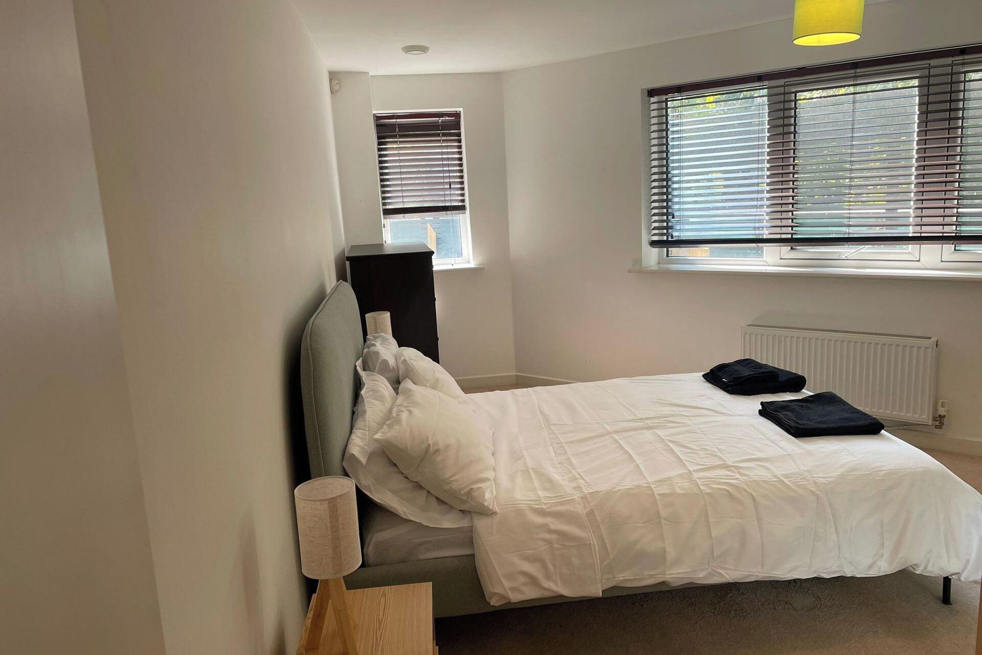 A bedroom with a bed, a bedside table, a lamp, a radiator, and a window with blinds.