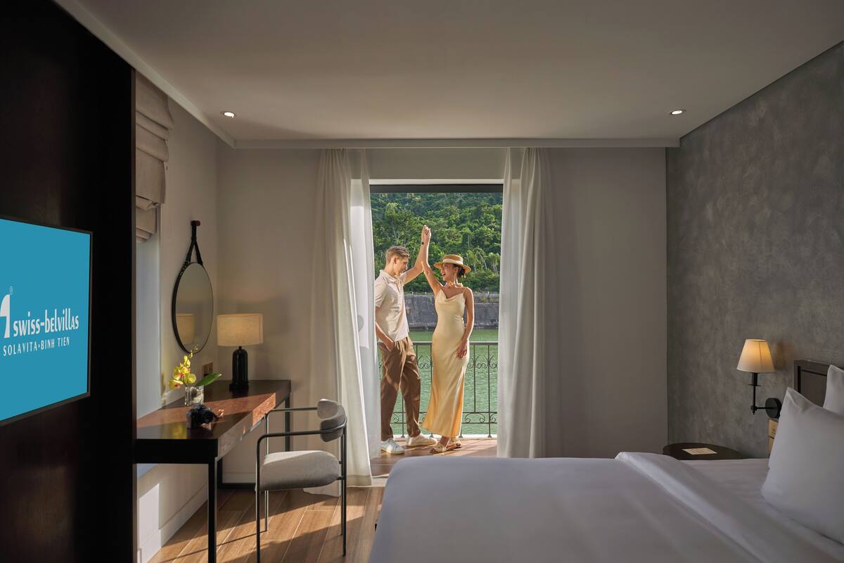 Une chambre d’hôtel comprenant un lit, un bureau avec un ordinateur, une chaise, une télévision et un balcon donnant sur un espace verdoyant.