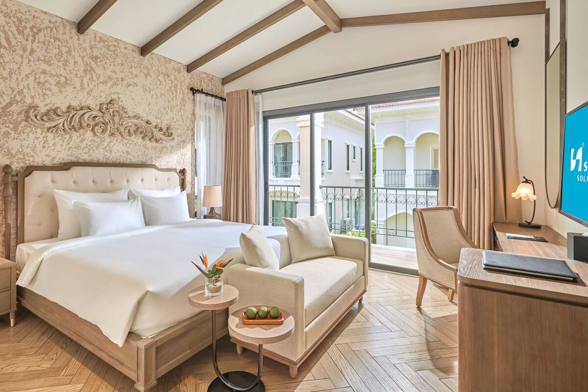 Une chambre d’hôtel avec un grand lit, un canapé, un bureau et une télévision. Elle dispose d’un balcon avec vue sur les immeubles.