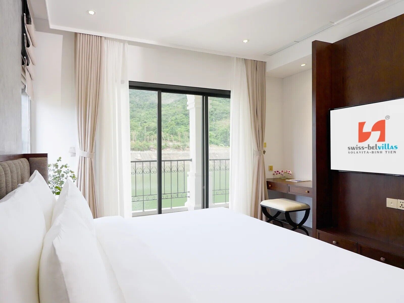 A hotel room with a large bed, a desk, a chair, and a balcony with a view of greenery.
