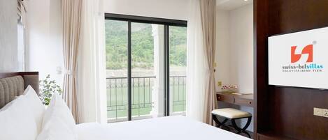 A hotel room with a large bed, a desk, a chair, and a balcony with a view of greenery.
