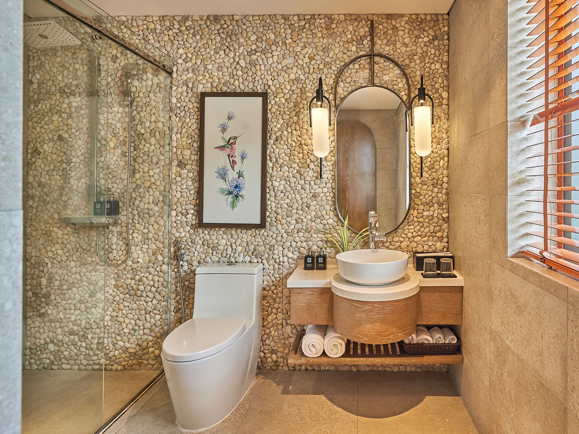 A modern bathroom with a stone wall, a glass shower enclosure, a toilet, a sink with a wooden vanity, and a mirror with wall-mounted lights.