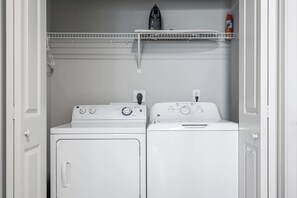 A laundry room with two stacked washers and dryers, a shelf above, and a power outlet on the wall.