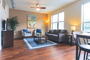 A living room with a brick wall, a ceiling fan, a poster, two armchairs, a sofa, a coffee table, and a dining table with chairs.