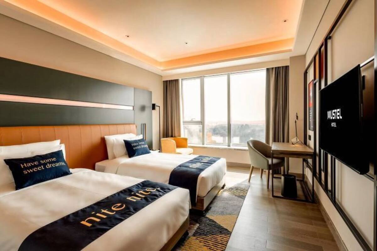 Ein Hotelzimmer mit zwei Betten, einem Schreibtisch, einem Stuhl, einem Fernseher und einem großen Fenster.