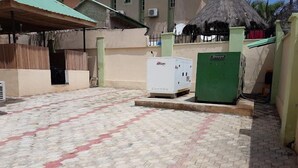 A paved courtyard with a thatched-roof structure, a white refrigerator, and a green utility box. - Solid Worth Hotels (Ilorin)