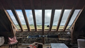 A rustic room with a stone wall and wooden beams, featuring a large window with a view of a rural landscape. - Le buron de Perrier  (Saint urcize)