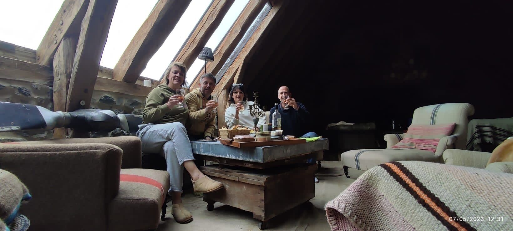 A cozy room with a wooden ceiling, a table with bottles, and a few people sitting and drinking.