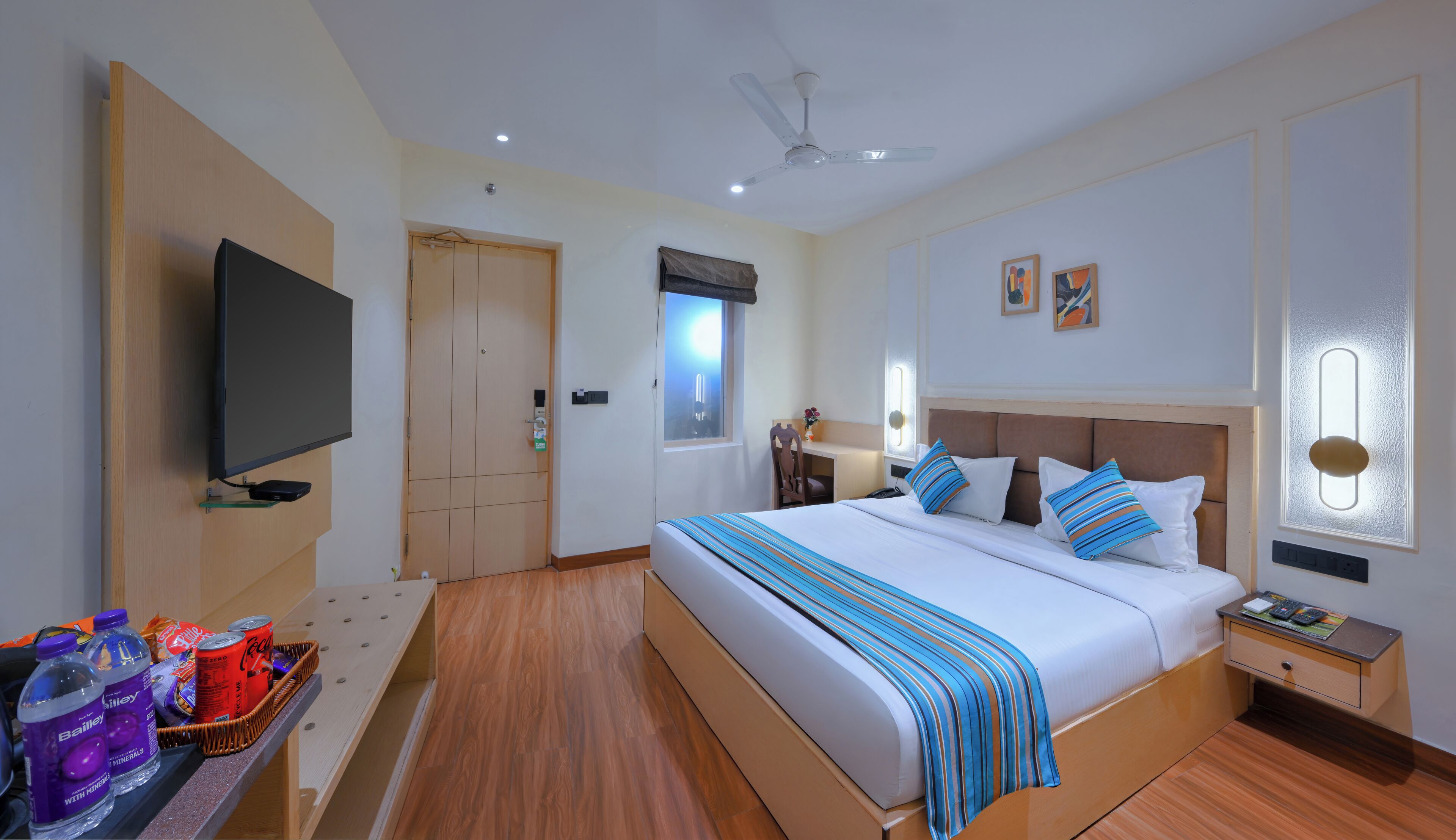 A hotel room with a bed, a TV, a desk with a basket of items, a window, and a ceiling fan.