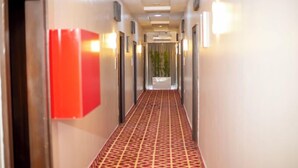 Un couloir d’hôtel avec un tapis à motifs, des luminaires fixés au mur et un objet rouge sur le mur de gauche.