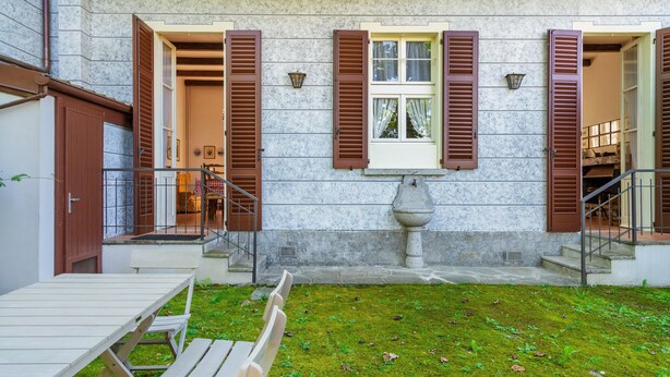 A building with a stone facade, wooden shutters, and a small garden with a table and chairs. - Holiday Home in Meina (Meina)