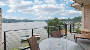 A balcony with a glass table and chairs, overlooking a lake with boats and houses in the background.