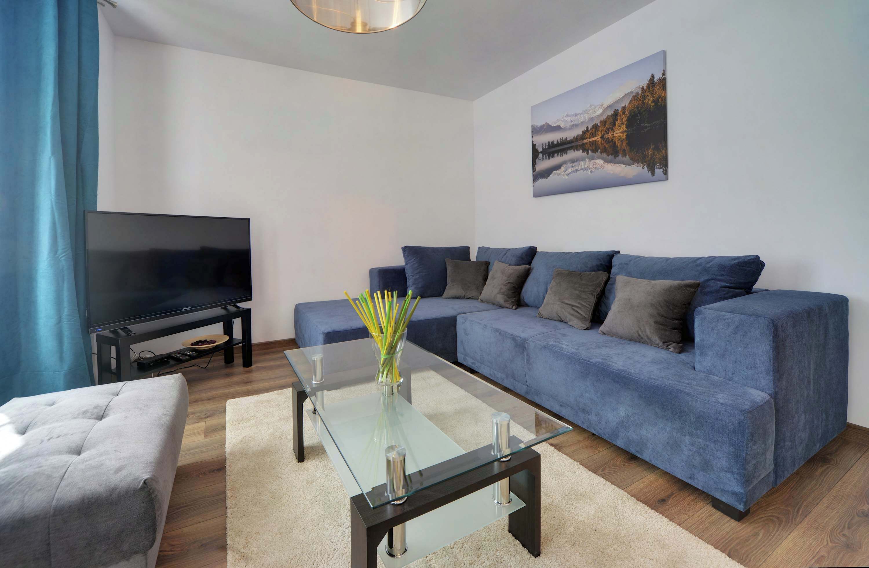 Una sala de estar moderna con un televisor de pantalla plana, un sofá azul, una mesa de centro de vidrio y un cuadro en la pared.