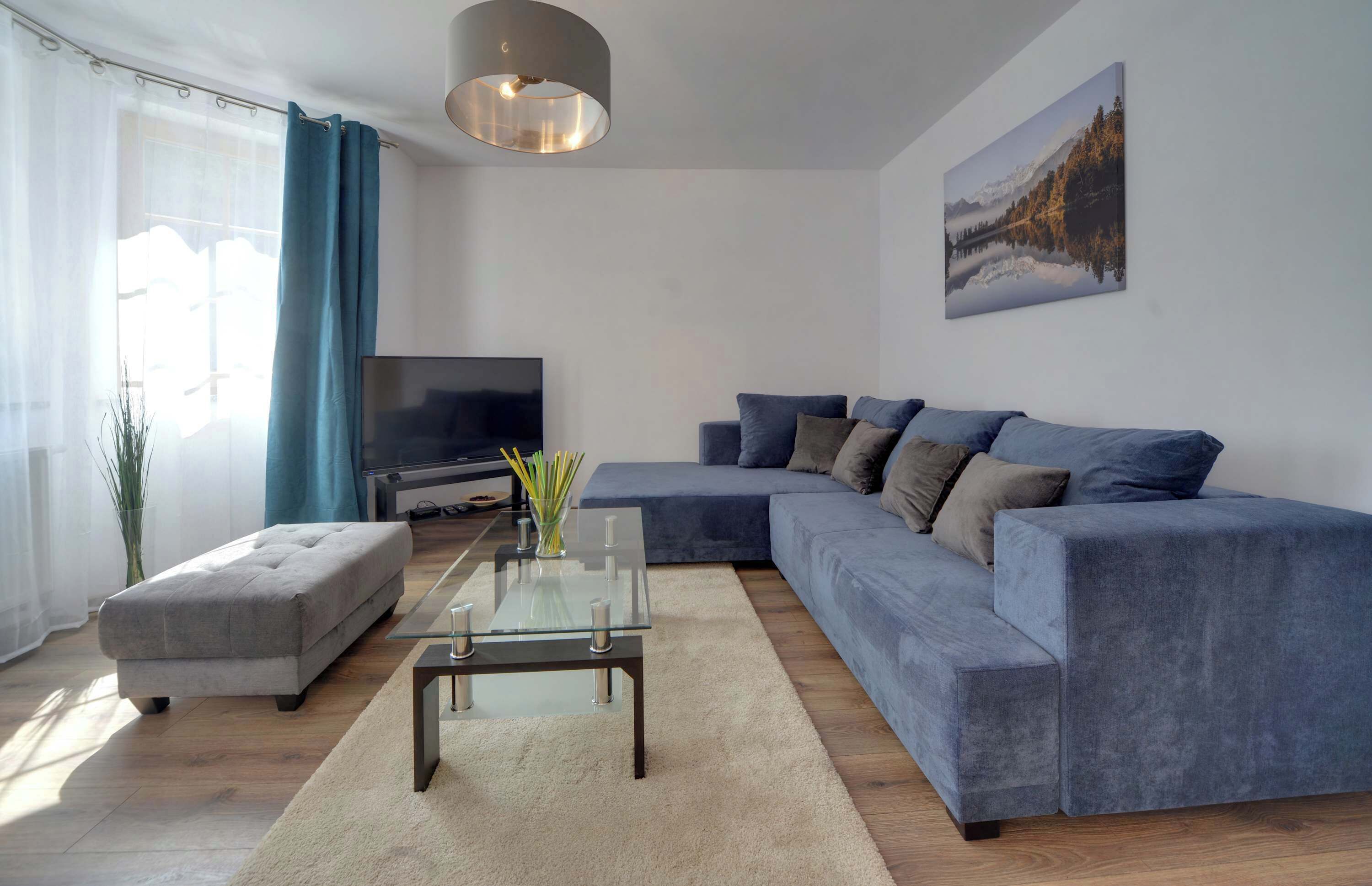 Ein modernes Wohnzimmer mit einem Ecksofa, einem Glastisch, einem Flachbildfernseher und einem großen Fenster mit Vorhängen.