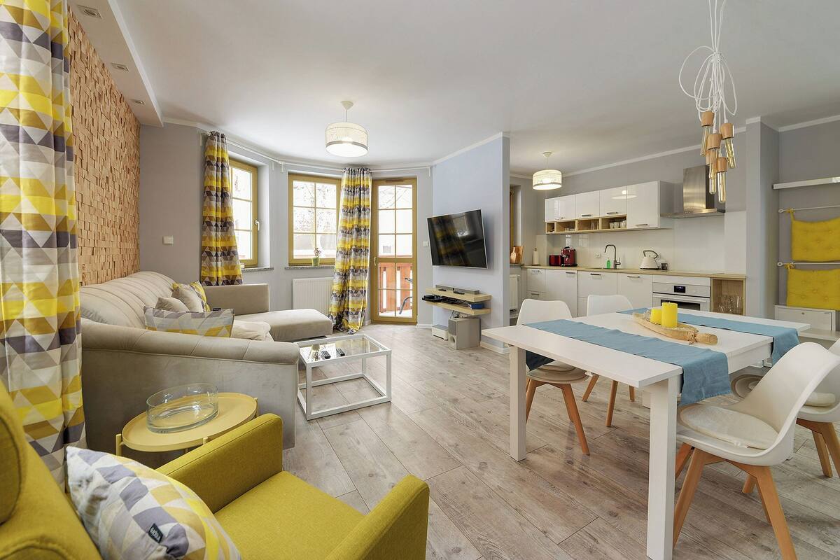 Un salon moderne avec un canapé, une table basse et des fauteuils jaunes. On y trouve également un coin repas avec une table et des chaises. Un téléviseur est fixé au mur, et on aperçoit une cuisine au fond de la pièce.