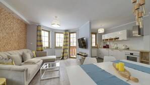 Een moderne woonkamer met een bank, salontafel en een eethoek met een tafel voor twee. Achtergrond toont een keuken met witte kasten en een televisie.