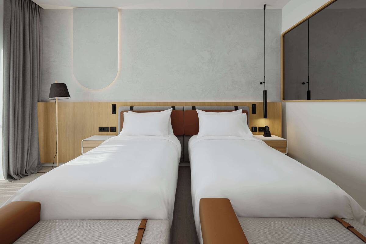 A modern hotel room with two single beds, a wooden headboard, a bedside table with a lamp, and a large mirror on the wall.
