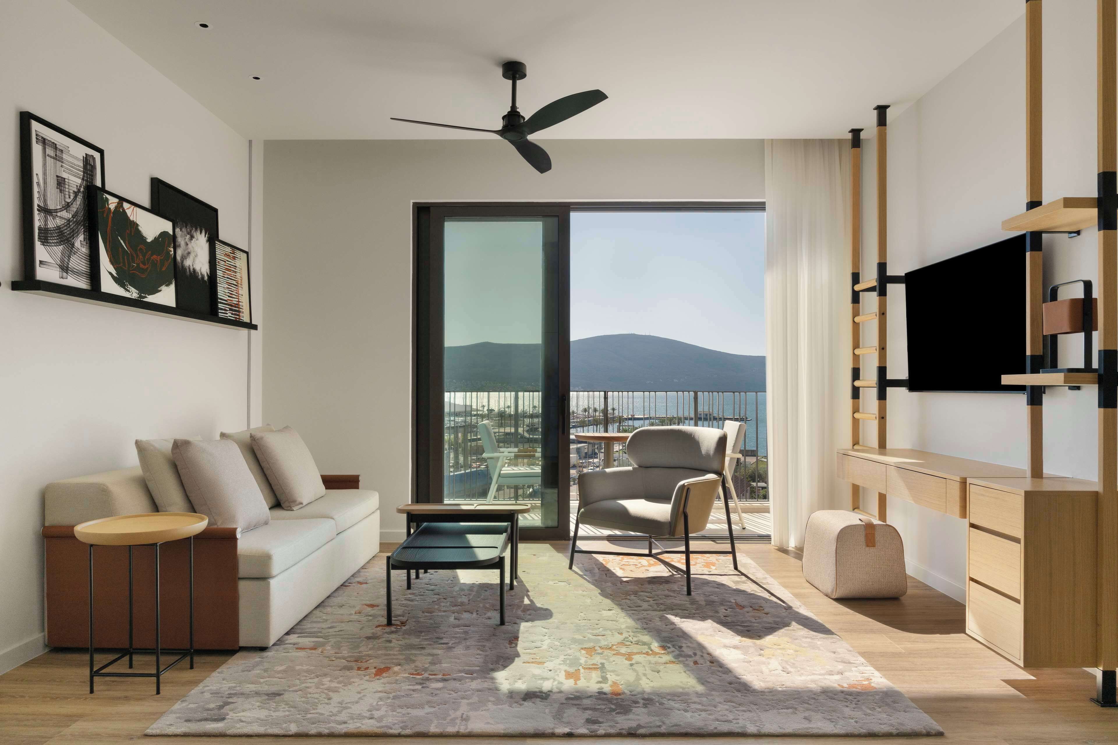 A modern living room with a sofa, coffee table, and armchair, a TV, and a balcony with a city view.
