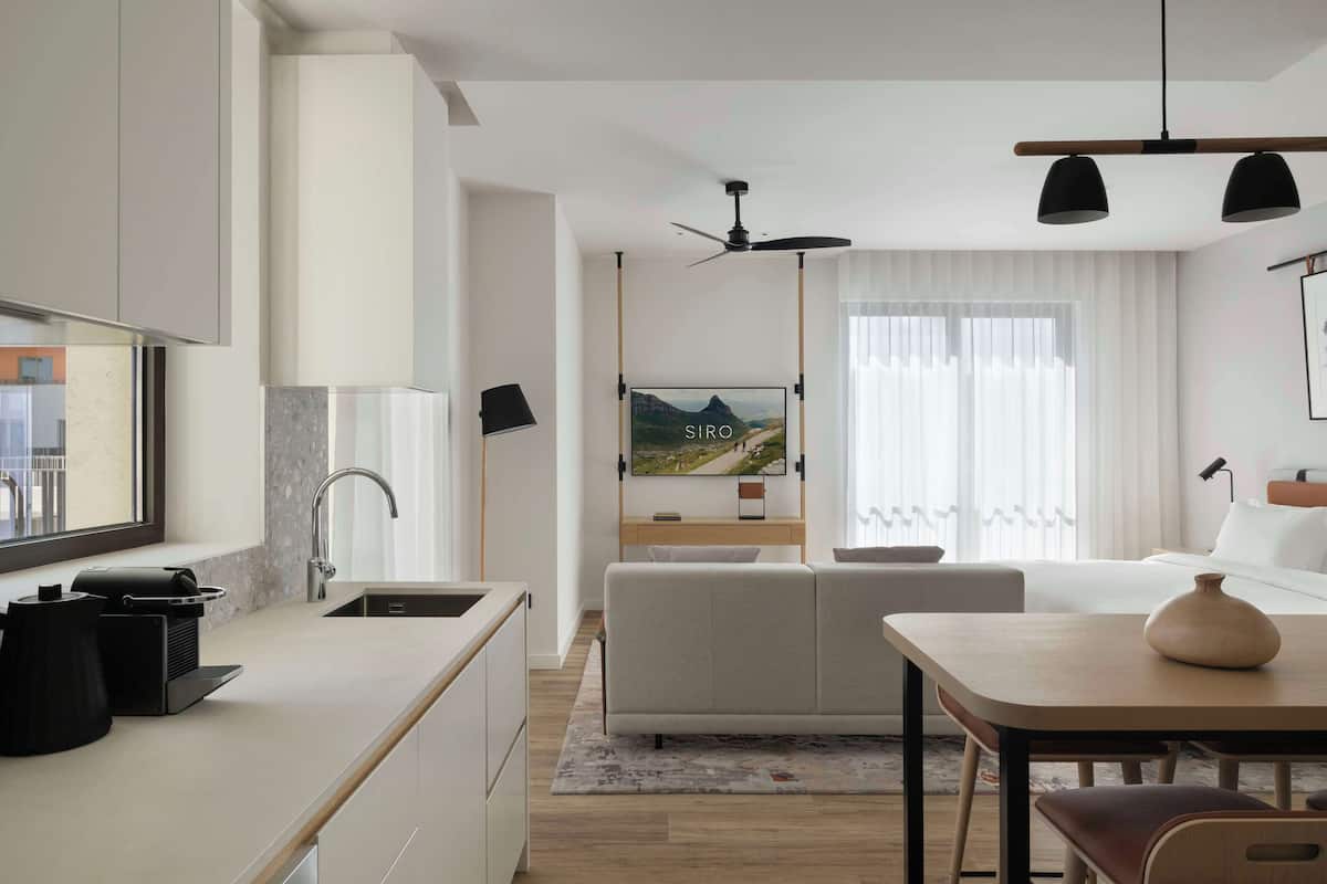 A modern hotel room with a kitchenette, a sofa, a dining table, and a large window with curtains.
