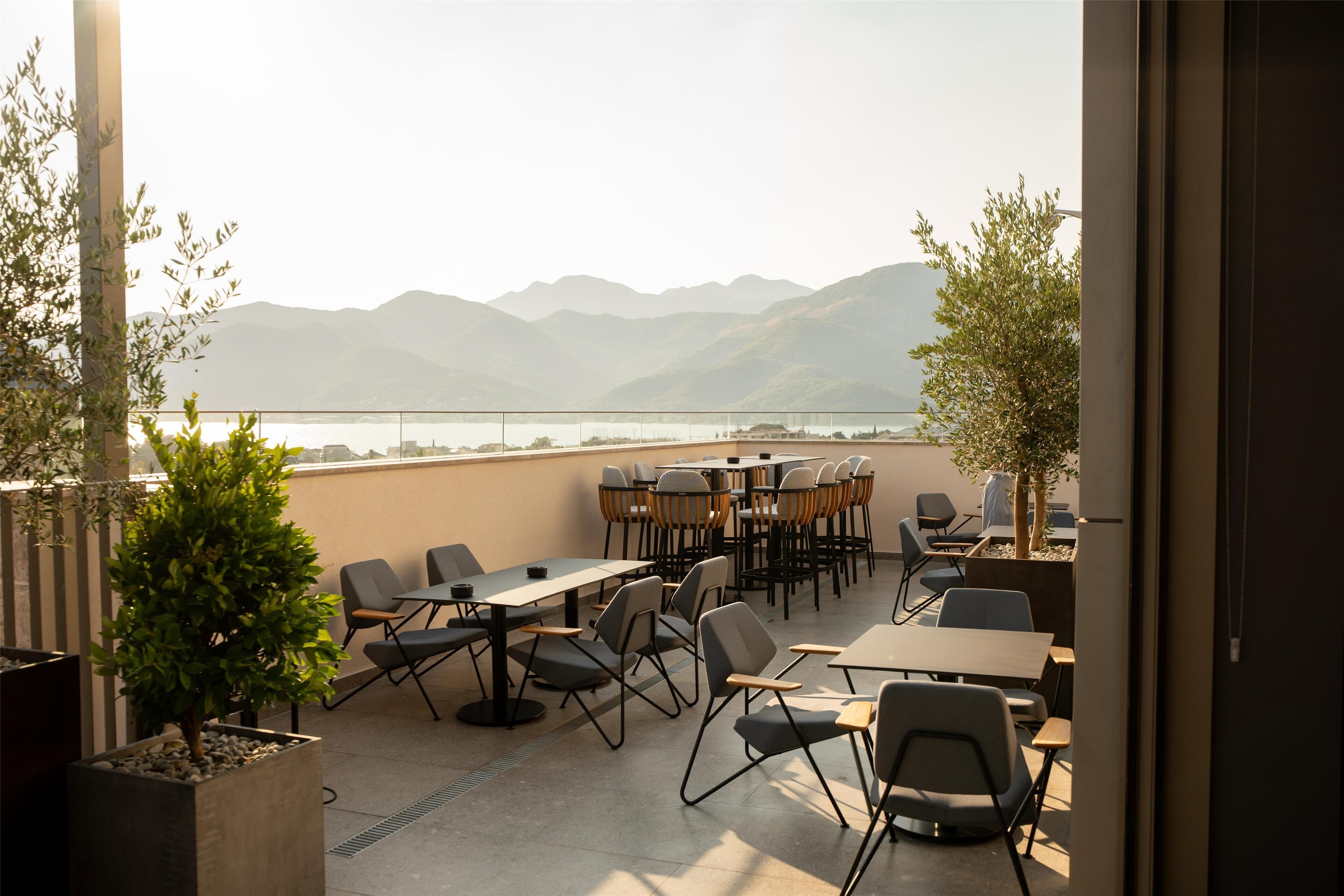 Terraza exterior con mesas y sillas, con vistas a un cuerpo de agua y montañas.